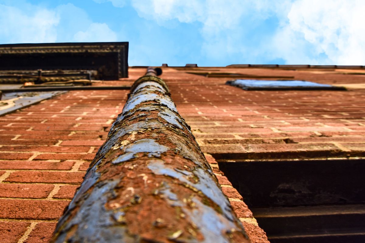 View of building exterior looking from the ground up to the sky showing a rusting gutter downpipe and broken masonry, mortar and brickwork. Image for this article highlights the risks that ageing facilities and infrastructure causes, including  structural deterioration to increased maintenance costs. 