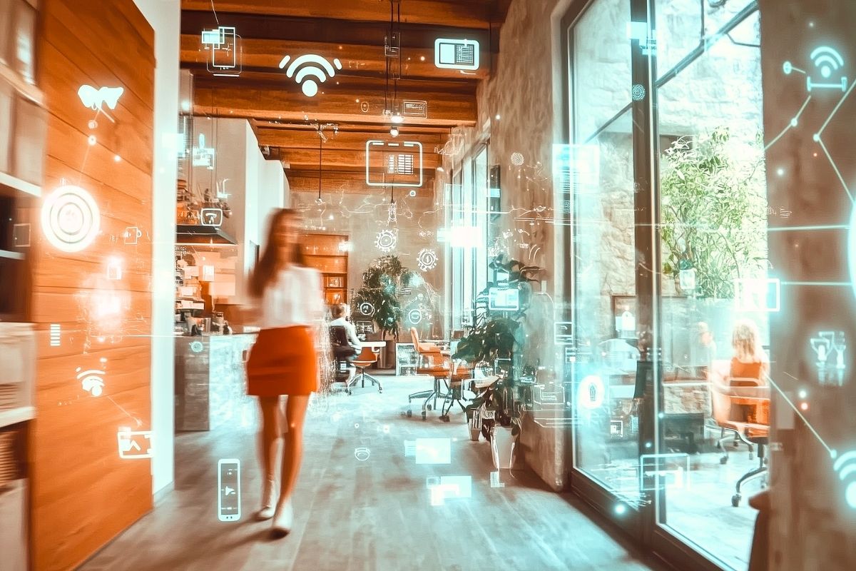 Modern office interior with Internet of Things (IoT) icons showing connected smart devices and equipment for facilities management, with office workers in the background and a person walking across the frame in motion blur.