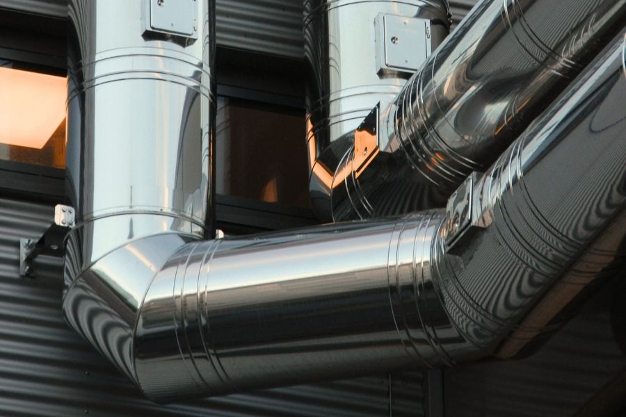 Close-up of industrial pipework with pressurised stainless steel ducts and connectors mounted on the exterior of a commercial building. The highly reflective pipes are part of a pressurised system designed for ventilation, heating, or air distribution in a factory or office facility.