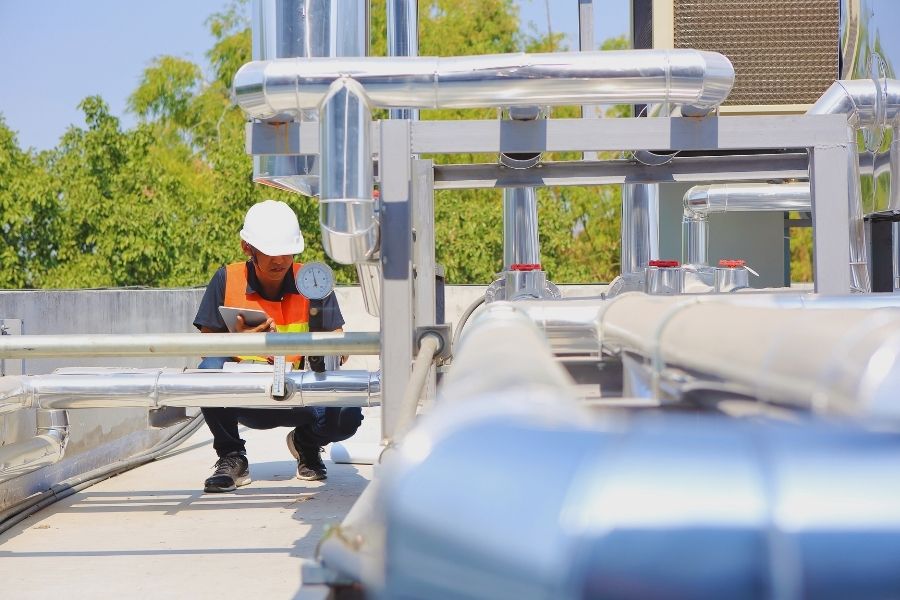 A system engineer wearing a white hard hat and orange safety vest is inspecting and recording pressure readings using a tablet. The engineer is surrounded by polished stainless steel pipework and pressure gauges on a rooftop facility, with greenery visible in the background.